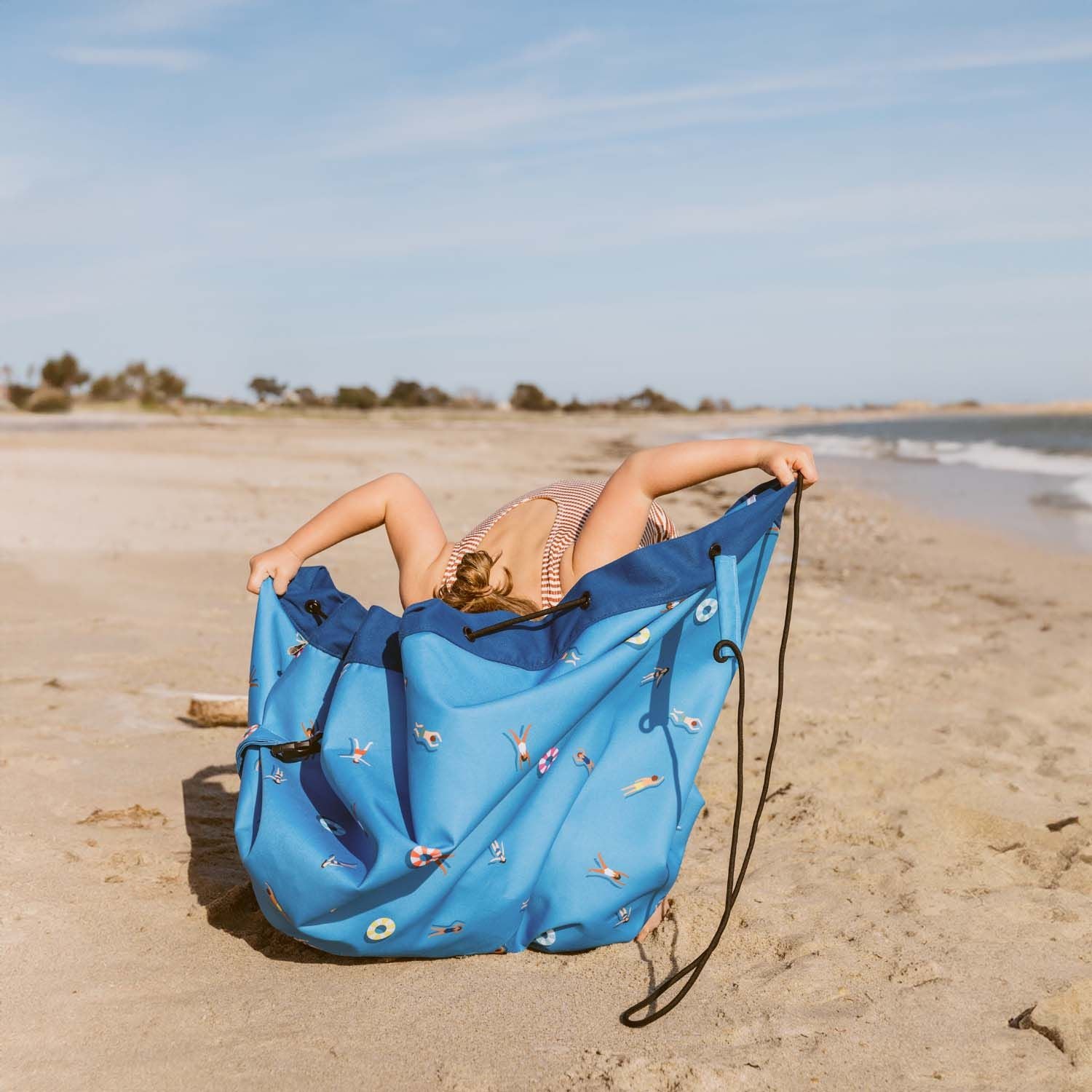swim fun outdoor beach bag, child is looking in the bag, standing at the beach, wearing a swim suit