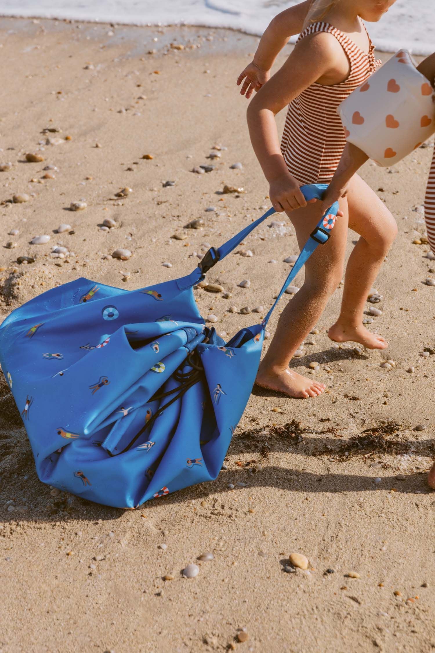 swim fun outdoor beach bag, children in swim suits are taking the bag by the shoulderstrap and they are pulling the bag over the beach