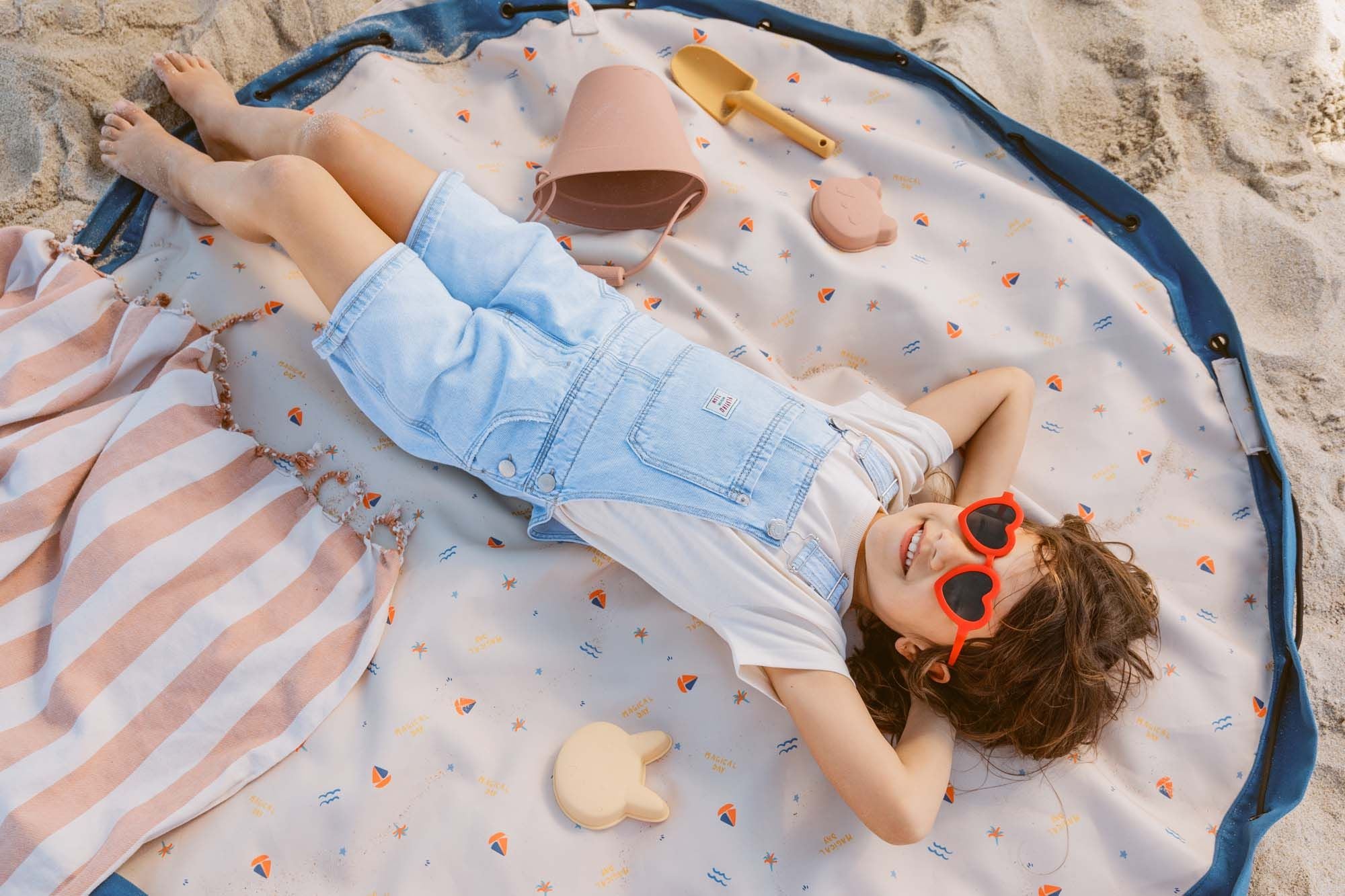 magical days outdoor beach bag, child is lying on the open mat, between beach toys,  funny sunglasses