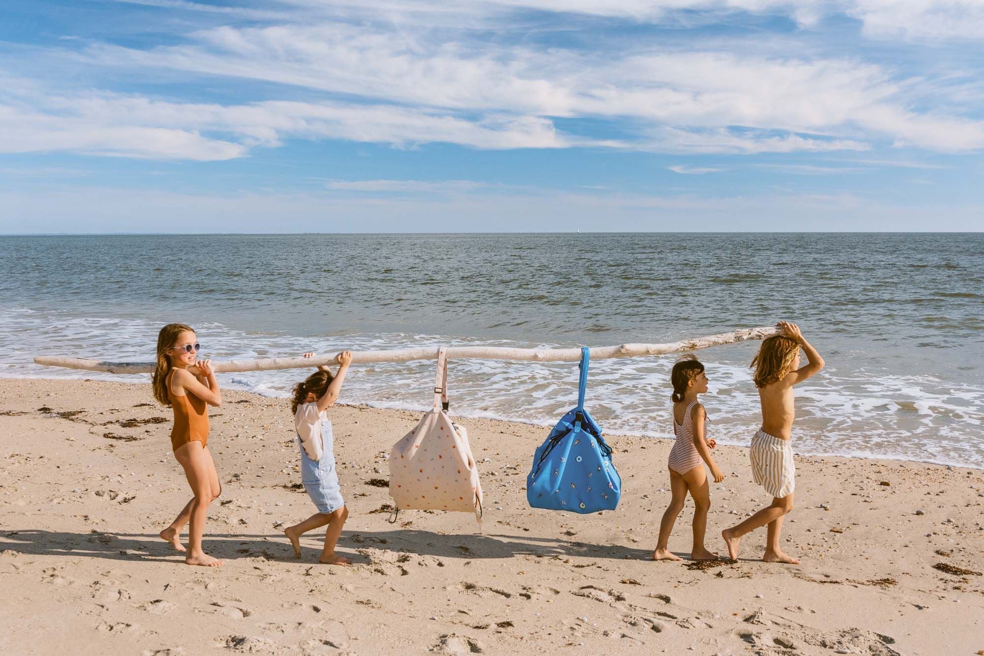 swim fun outdoor beach bag, children are carrying a wooden stick with the beach bags hanging on it.