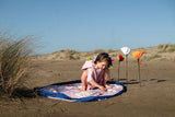 Beach outdoor storage bag, child sitting on a beach mat with flowers and a book, surrounded by sand dunes.