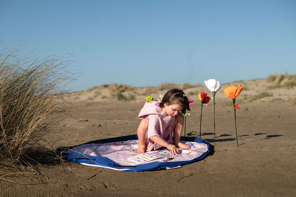 Beach outdoor storage bag, child sitting on a beach mat with flowers and a book, surrounded by sand dunes.