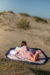 Beach outdoor storage bag, person sitting on a colorful mat on a sandy beach with grasses, reading a book