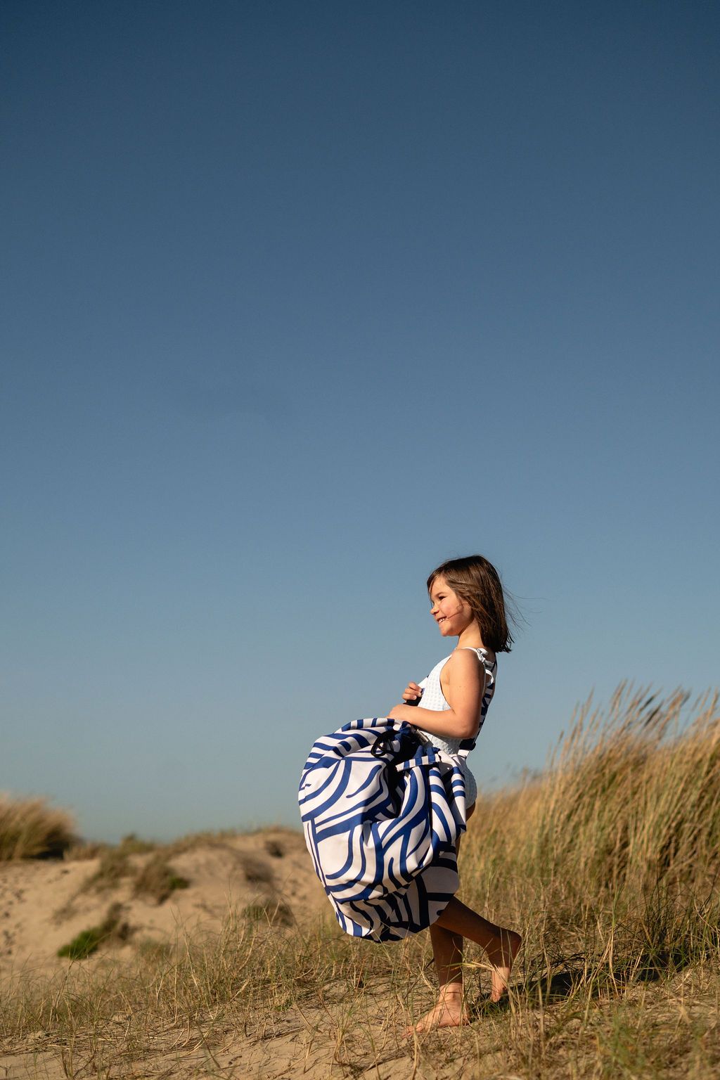 pirate outdoor playmat, child in a blue and white swimsuit standing in a field of dunegrass with a clear blue sky, carrying the bag