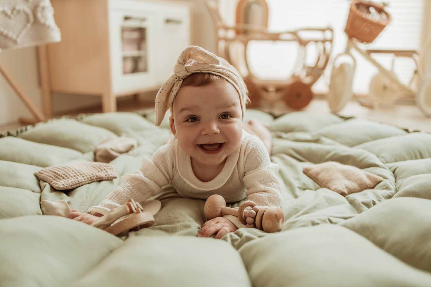 Bloom meadow green organic babymat, baby lying on open mat between toys, laughing, wooden kitchen and tricycle