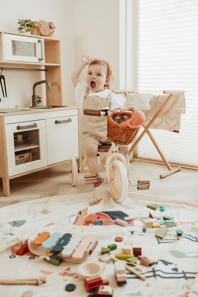 farm toy storage bag and play mat, child riding on tricycle and waving, lots of wooden toys and wooden play kitchen