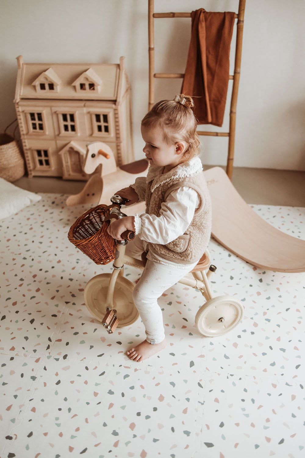 child riding with tricycle on terrazzo puzzlemat, between wooden toys such as rocking horse and slide